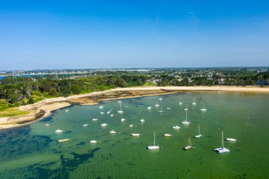 Anse de Trehennarvour, Saint Philibert 'te kumlu bir sahil ve yemyeşil bir orman yakınlarındaki sığ zümrüt suya demirlemiş sayısız teknenin hava görüntüsü..