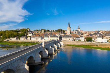 Fransa 'nın orta kesimindeki La Charite sur Loire' da klasik nehir kenarı manzarası. Loire Nehri üzerinde taş kemer köprüsü, pastel konaklar ve derin mavi gökyüzünün altında önemli bir kilise kulesi bulunuyor. Ortaçağ mimarisi ve sakin su,