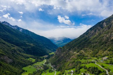 Manzaralı bir dağ yolu, İtalya 'nın Val dAoste kentindeki orman yamaçları ve uzak karlı zirvelerle çevrili yemyeşil bir vadi boyunca kıvrılır. Manzara canlı çayırlara, çarpıcı yüksekliğe ve parlak bir ışık ve bulutların karışımına sahiptir.