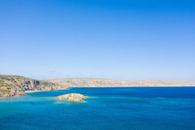 Girit 'teki Vai sahilinin engebeli kıyı şeridine yakın küçük kayalık bir adacığı çevreleyen berrak turkuaz suları. Güneşli manzarada kurak tepeler ve canlı mavi bir gökyüzü, sakin bir Akdeniz manzarası yaratıyor..