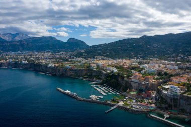 İtalya 'nın Sorrento kentinin yüksek açılı manzarasında canlı bir marina ve derin mavi Akdeniz sularının üzerindeki dramatik uçurumlara tünemiş renkli binalar görülüyor. Sahne engebeli dağlar ve dokulu bulutlarla dolu bir gökyüzü üzerine kurulmuştur..