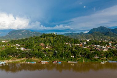 Mekong Nehri 'ne bakan yeşil bir tepeyi ve Luang Prabang, Laos' un çatılarını altın bir stupa taçlandırır. Ahşap tekneler, uzak dağlar ve dramatik bulutlarla dolu canlı mavi bir gökyüzünün altındaki çamurlu nehir boyunca uzanır..