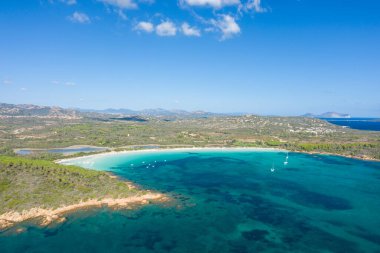 Sardinya 'daki Cala Brandinchi' nin geniş hava manzarası, derin mavi su, demirli yelkenliler ve uzak dağlarla dolu açık bir gökyüzü altında yemyeşil bir kıyı şeridi barındırıyor..