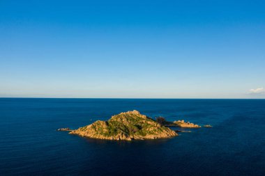 Yeşil bitki örtüsüyle kaplı küçük kayalık bir ada, Sardunya 'nın Lotzorai kenti yakınlarındaki derin mavi Akdeniz sularıyla çevrili sıcak altın güneş ışığıyla aydınlanır. Açık gökyüzü ve sakin deniz huzurlu ve geniş bir atmosfer yaratır..
