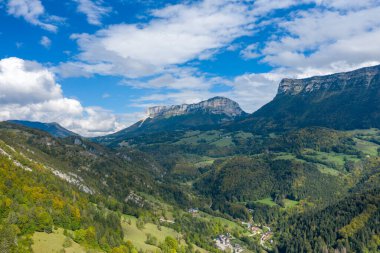 Chartreuse Massif 'in geniş hava perspektifi çarpıcı kireçtaşı uçurumları, yuvarlanan yeşil vadiler ve dağınık bulutlarla dolu canlı mavi bir gökyüzünün altında dağılmış orman yamaçlarını gözler önüne serer. Sahne doğal hatları ve yemyeşil bitki örtüsünü yakalar.