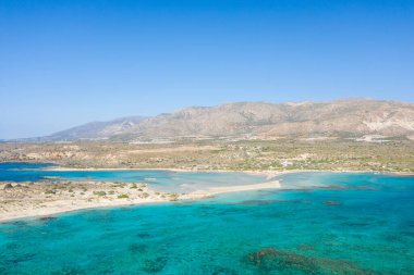 Yunanistan 'ın Girit kentindeki Elafonisi plajının hava perspektifi, çarpıcı turkuaz sular, kumlu bir yarımada ve kusursuz mavi gökyüzünün altında kurak bir dağ zemini sergiliyor. Sahne açık, güneşli ve doğal olarak huzurlu..