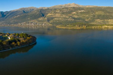 Ioannina Gölü 'ne uzanan ağaçlarla kaplı bir yarımadanın havadan görünüşü, sakin su Pindus dağlarının engebeli yamaçlarını yansıtıyor. Sahnede gün ışığı, doğal dokular ve Yunanistan 'ın Epirus kentinde barışçıl bir göl manzarası yer alıyor..