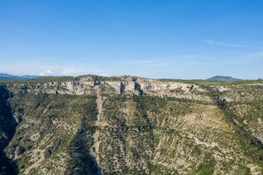 Navacelles Sirki 'ndeki engebeli, yeşil noktalı yamaçlı dramatik kireçtaşı kanyonun yüksek açılı perspektifi. Açık mavi gökyüzü ve güneş ışığı kayalık arazinin dokularını ve hatlarını vurgular..