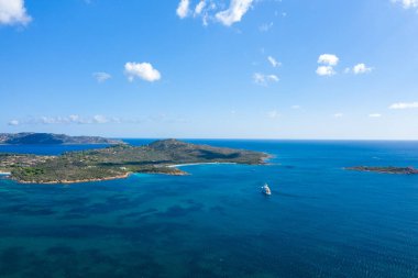 Spiaggia la Salinedda, Sardinya 'nın engebeli kıyı şeridine demirlemiş bir yatın geniş bir hava perspektifi. Derin mavi su ve parlak gökyüzü altında dağınık bulutlar..