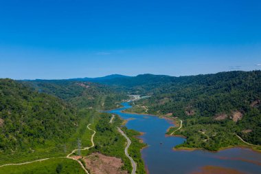Hava görüntüsü, bereketli, ormanlık tepeler ve parlak mavi gökyüzünün altındaki dağınık açıklıklar arasında akan dolambaçlı bir nehir olduğunu gösteriyor. Manzara doğal kıvrımlar, yoğun bitki örtüsü ve açık suları barındırıyor, bir huzur hissi yaratıyor ve bozulmamış.