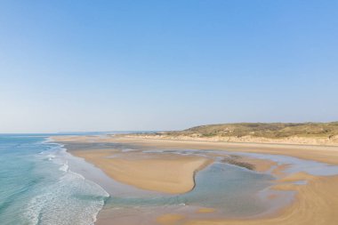 Plage de la Vieille eglise 'de dalgalı dalgalar ve dalgalı dalgalar içeren sakin bir kumlu sahil şeridi vardır. Arka planda alçak çimenli kum tepeleri yükseliyor engin, berrak mavi gökyüzünün altında.