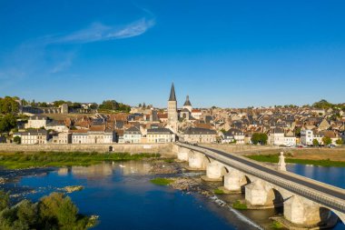 La Charite sur Loire 'deki tarihi taş köprü ve pastel evler altın ışıkla yıkanırken, manastır şehrin üzerinde yükseliyor. Loire nehri canlı çatıları ve berrak mavi gökyüzünü yansıtıyor. Huzurlu bir Loire Valley atmosferini çağrıştırıyor..