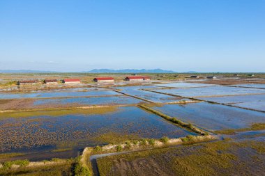 Kamboçya, Kamboçya yakınlarındaki geniş su basmış tuz buharlaştırma göletlerinin kenarında kırmızı çatıları olan kırsal tuz depolama barakaları. Manzara geometrik su desenleri, açık gökyüzü ve uzak mavi dağlar içerir, sakin atmosferi yakalar.