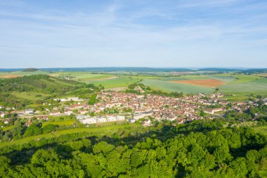 Yuvarlanan yeşil tarlalar ve ormanlık araziler arasında uzanan Varzy köyünün hava manzarası, kırmızı kiremitli çatılar ve açık mavi gökyüzünün altında uzanan yamalı tarlalar..