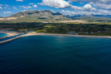 Kumlu sahil ve dalgakıranla buluşan açık mavi deniz manzarası, yemyeşil kırsal ve engebeli dağlar Orosei, Sardunya yakınlarındaki dağınık bulutların altında.