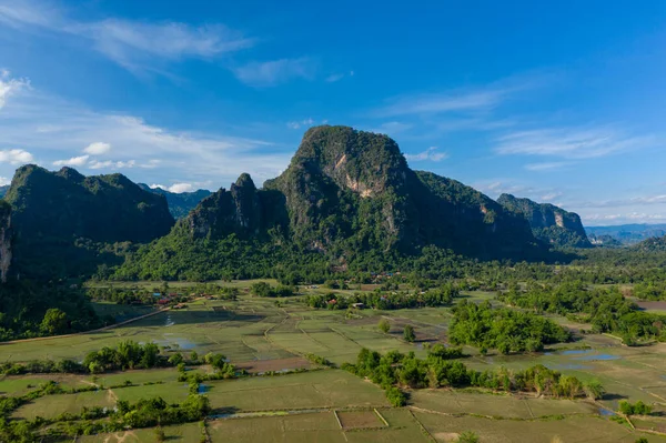 Dramatik karst dağları yeşil tarlaların ve Laos kırsalındaki dağınık su havuzlarının üzerinde yükseliyor. Açık arazide canlı mavi gökyüzü, yemyeşil bitki örtüsü ve Güneydoğu Asya 'nın tipik doğal desenleri yer alıyor..