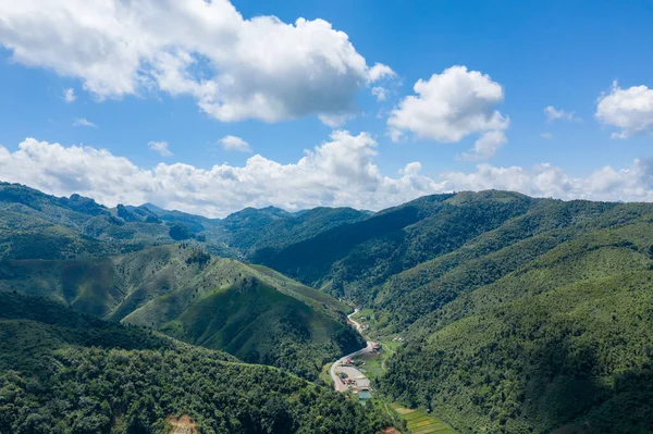 Dolambaçlı bir yolu olan yemyeşil bir vadinin geniş manzarası ve canlı mavi bir gökyüzünün altında dağılmış kırsal binalar ve Phou Khoun ile Luang Prabang, Laos arasındaki yolda tüylü bulutlar. Manzara katmanlı