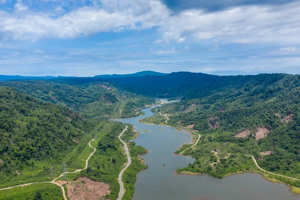 Bereketli, yoğun ormanlık tepeler ve parlak bir gökyüzünün altında berrak araziler arasında dolaşan bir nehrin havadan bakış açısı. Manzara uzak bir kırsal bölgede dolambaçlı su, canlı yeşillik ve doğal kıvrımlara sahiptir..