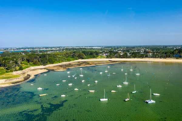 Anse de Trehennarvour, Saint Philibert 'te kumlu bir sahil ve yemyeşil bir orman yakınlarındaki sığ zümrüt suya demirlemiş sayısız teknenin hava görüntüsü..