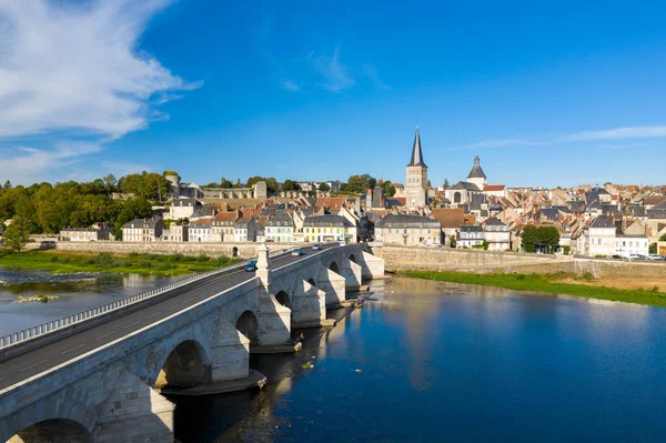 Fransa 'nın orta kesimindeki La Charite sur Loire' da klasik nehir kenarı manzarası. Loire Nehri üzerinde taş kemer köprüsü, pastel konaklar ve derin mavi gökyüzünün altında önemli bir kilise kulesi bulunuyor. Ortaçağ mimarisi ve sakin su,