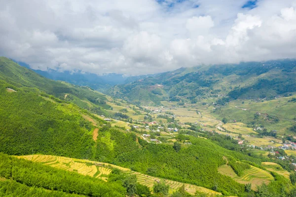 Sapa, Vietnam yakınlarındaki kırsal bir vadiyi çevreleyen yemyeşil tepeler ve katmanlı pirinç teraslarının geniş panoramik manzarası. Bulutlarla dolu gökyüzü ışıl ışıl manzarayı ve uzak köyü aydınlatıyor..
