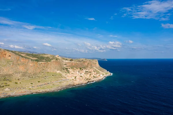 Monemvasias 'ın derin mavi Akdeniz' le buluşan dramatik uçurumlarının hava görüntüsü. Canlı bir gökyüzünün altında kayalık kıyı şeridine kurulmuş tarihi bir köy. Sahnede desenli taş, yemyeşil ve geniş su yer alıyor..
