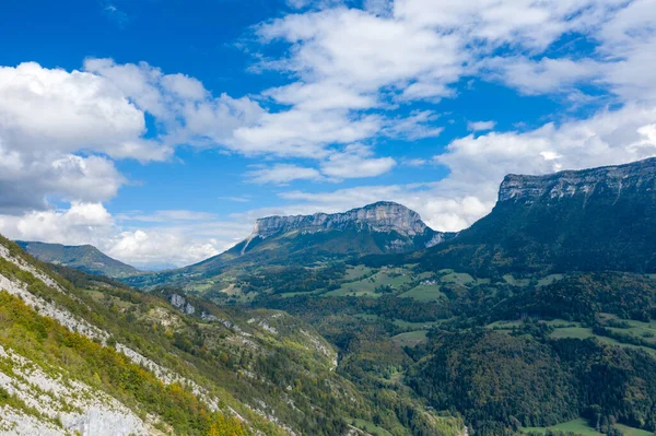 Chartreuse Massif 'teki dramatik kireçtaşı kayalıkları ve orman yamaçlarının geniş bir manzarası var. Dağınık bulutlar ve aşağıda canlı yeşil vadiler var. Sahne, bu ikonik alp bölgesinin engebeli arazisini ve doğal güzelliğini vurguluyor..