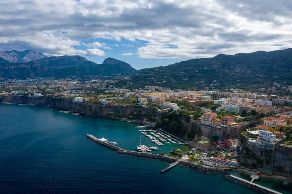 İtalya 'nın Sorrento kentinin yüksek açılı manzarasında canlı bir marina ve derin mavi Akdeniz sularının üzerindeki dramatik uçurumlara tünemiş renkli binalar görülüyor. Sahne engebeli dağlar ve dokulu bulutlarla dolu bir gökyüzü üzerine kurulmuştur..