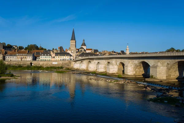 La Charite sur Loire 'ın ikonik taş köprüsü, ortaçağ manastır kulesi ve nehir kıyısındaki sakin Loire nehrine yansıyan mavi gökyüzü manzaralı geniş manzarası. Sahne, şehirlerin mimari mirası ve sükuneti ön plana çıkarıyor.