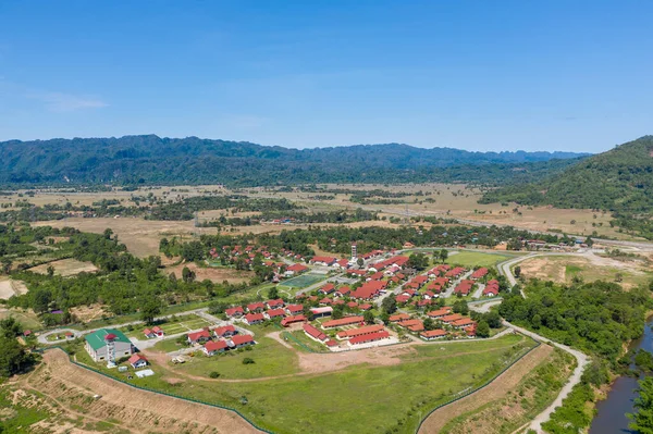 Geniş hava perspektifi, açık araziler, dağınık ağaçlar ve açık mavi gökyüzünün altındaki uzak ormanlık tepelerle çevrili kırmızı çatılı binalarla küçük bir yerleşim yeri yakalar. Manzara gelişmiş bölgelerle doğal bölgeler arasındaki zıtlığı vurgular.