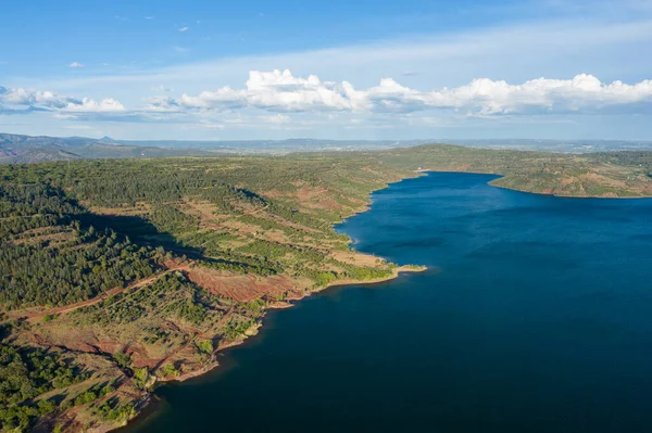 Yoğun bir orman ve kızıl toprağın, Salagou Gölü 'nün canlı mavi sularıyla buluşup dağınık bulutlarla kaplı bir gökyüzünün altında uçarak geçen bir kıyı şeridi..