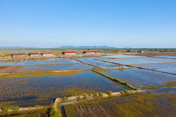 Kamboçya, Kamboçya yakınlarındaki geniş su basmış tuz buharlaştırma göletlerinin kenarında kırmızı çatıları olan kırsal tuz depolama barakaları. Manzara geometrik su desenleri, açık gökyüzü ve uzak mavi dağlar içerir, sakin atmosferi yakalar.