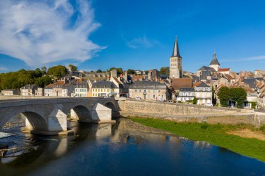 Fransa 'nın orta kesimindeki La Charite sur Loire' ın klasik nehir kenarı manzarası, Loire Nehri üzerinde tarihi bir taş köprü, pastel renkli evler ve canlı mavi gökyüzünün altında uzun bir kilise kulesi. Sahne ortaçağ mimarisini harmanlıyor.