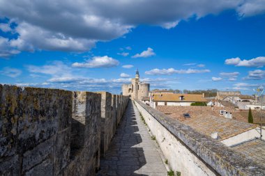 Tarihi duvarlar boyunca uzanan düz taş patikalar, dokulu çatılar ve dağınık bulutlarla dolu canlı mavi bir gökyüzü tarafından çerçevelenmiş Aigues-Mortes 'in önde gelen ortaçağ kulesine götürür..