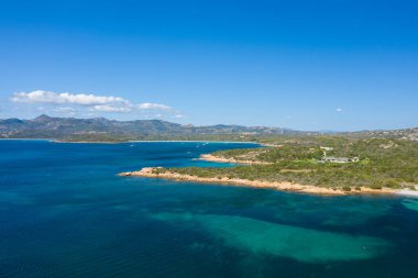 Sardunya 'daki engebeli bir yarımadanın havadan görünüşü canlı mavi Akdeniz sularıyla çevrili, uzak tepeler ve parlak bir gökyüzü altında dağınık bulutlar..
