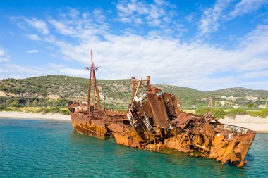 Paslanmış Dimitrios gemi enkazı, kısmen kumlu bir Yunan kıyı şeridinde, parlak mavi bir gökyüzünün altında yeşil tepeler ve dağınık evler altında, berrak turkuaz sulara batmış vaziyette bulunuyor..