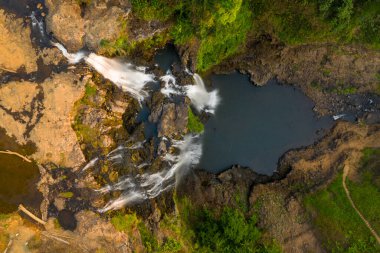 Tad Yuang şelalesinin yukarıdan aşağıya doğru görüntüsü, Güney Laos 'taki Bolaven Platosu' ndaki yemyeşil bitki örtüsü ve topraksı araziyle çevrili, engebeli bazalt kayaların üzerinden çağlayarak geçen karanlık bir havuzu yakalar. Sıcak güneş ışığı kayanın dokusunu güçlendirir.