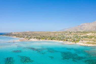 Yunanistan 'ın Girit kentindeki Elafonisi plajının geniş hava manzarası, berrak turkuaz su, kumlu bir kıyı şeridi ve bulutsuz mavi gökyüzünün altındaki engebeli tepeleri gösteriyor. Sahne geniş, güneşli ve doğal olarak canlı görünüyor..