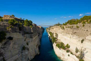Açık mavi su, sarp, yeşil çam ağaçlarıyla kaplı güneş ışığında kireçtaşı duvarlarla çevrili dar Corinth Kanalı 'ndan akar. Kavisli bir köprü, çarpıcı bir manzara yaratan, canlı bir gökyüzünün altındaki dramatik geçidi kaplar..