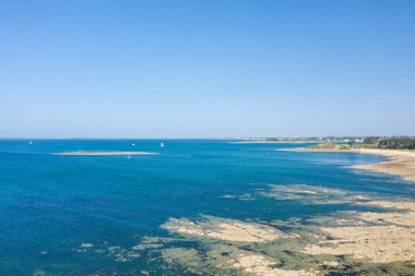 Kayalık sığ suların, canlı turkuvaz suların ve Saint Philibert yakınlarındaki Anse de Trehennarvour yakınlarındaki bulutsuz mavi gökyüzünün altında kumlu bir kıyı şeridinin geniş bir görüntüsü..