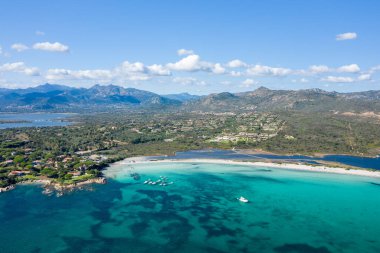 Sardunya 'daki Spiaggia Lu Impostu' nun havadan görünüşü. Parlak turkuaz deniz, beyaz kumlu sahil şeridi ve dağınık bulutlu bulutlu, açık bir gökyüzü altında yemyeşil dağlar..
