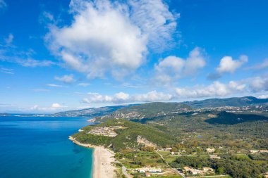 Yunanistan 'ın Epirus kentindeki Karavostasi kıyı şeridinin hava manzarası, turkuaz deniz, kumlu plaj, ormanlık bir tepenin üzerinde modern bir tatil beldesi ve dağınık bulutlu parlak bir gökyüzü altında geniş yeşil dağlar yer alıyor. Akdeniz manzarası...