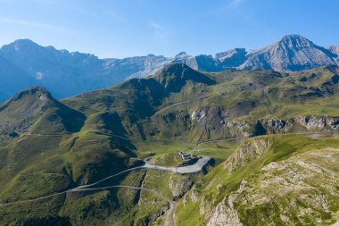 Gavarnie Gedre yakınlarındaki Pireneler 'de yeşil yamaçları ve engebeli araziyi geçen eğimli bir dağ yolunun hava manzarası. Güneşli tepeler ve dramatik kayalık tepeler berrak mavi gökyüzünün altında yükselir..