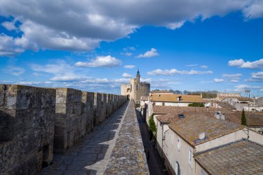 Düz taş duvar Aigues-Mortes 'te tarihi bir kuleye doğru uzanır, dokulu çatıları ve canlı mavi gökyüzü altında dramatik bulutlar.