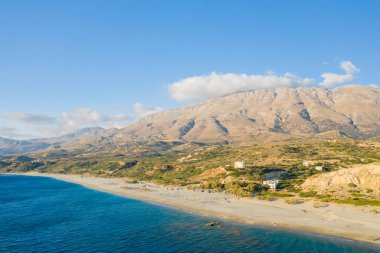 Geniş kumlu kıyı şeridi Triopetra, Girit kıyıları boyunca uzanan derin mavi denizle güneşli engebeli dağlarla ve açık gökyüzü altında dağınık evlerle birleşiyor..