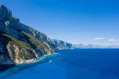 Cala Goloritze, Baunei, Sardinya yakınlarındaki parlak mavi Akdeniz boyunca yeşil bitki örtüsüyle kaplı yüksek kireçtaşı kayalıkları. Canlı kıyı manzarası berrak bir gökyüzünün altında parlak güneş ışığıyla yıkanır..