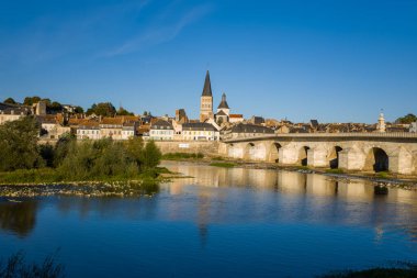 Akşam güneşi, tarihi taş köprüye, manastır kulesine ve La Charite sur Loire 'nin pastel nehir kenarındaki evlerine sıcak bir ışık saçıyor. Loire Nehri, ortaçağ mimarisi ve mavi gökyüzünü yansıtarak sakin bir Loire Vadisi sahnesi oluşturur..