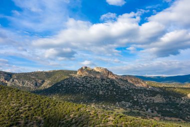 Dağınık bitki örtüsü ve gökyüzünde dramatik bulutlarla yuvarlanan yeşil tepelerin üzerinde engebeli bir kayalık yükseliyor. Manzara yumuşak gün ışığında yıkanır, açıklık ve sükunet hissi yaratır..