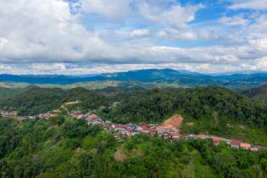 Yeşil tepeler ve dağınık bulutlarla dolu gökyüzünün altında uzanan uzak mavi dağlarla dolu yemyeşil bir orman sırtı boyunca tünemiş bir köyün havadan bakış açısı. Manzara doğal hatları, yoğun bitki örtüsünü ve