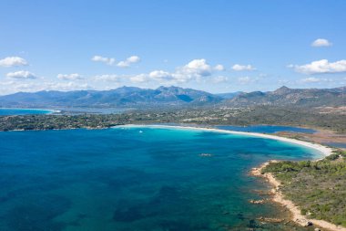 Sardunya 'daki Spiaggia Lu Impostu' nun hava perspektifi. Beyaz kum, turkuaz su ve engebeli dağlar ve dağınık bulutlar tarafından çerçevelenmiş yemyeşil bir göl..